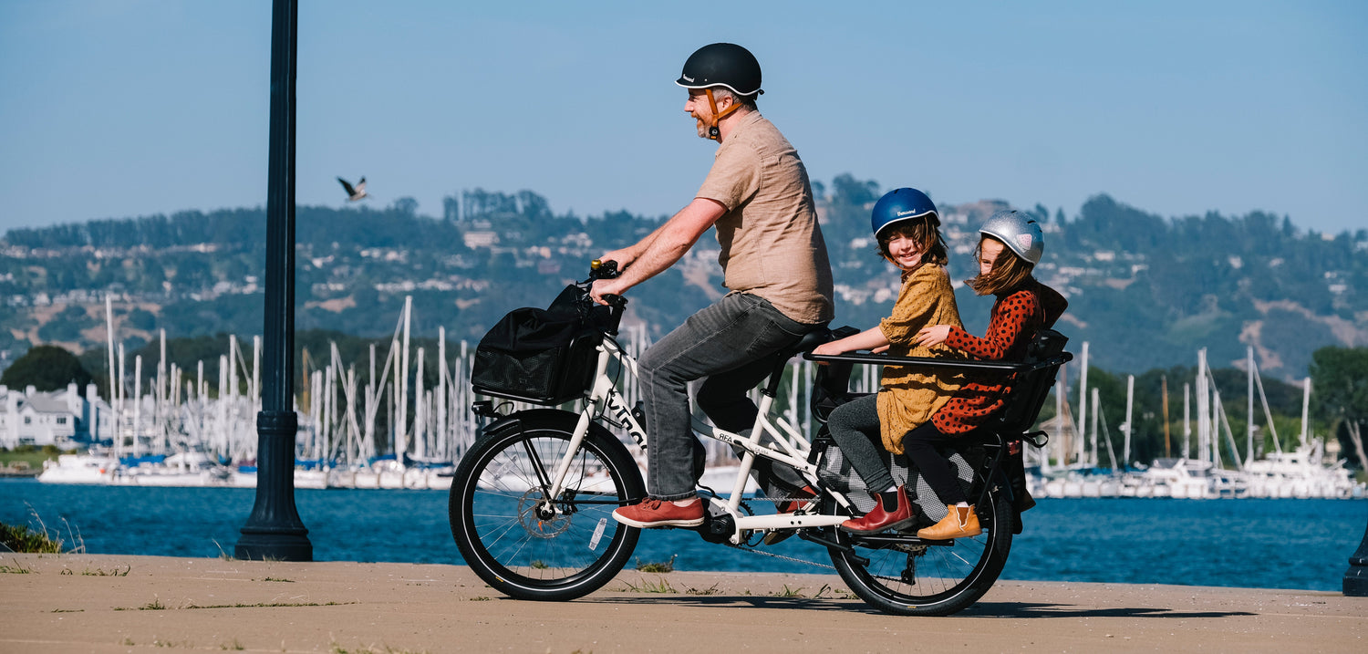 Family of three riding a cargo bike by a waterfront with sailboats in the background.